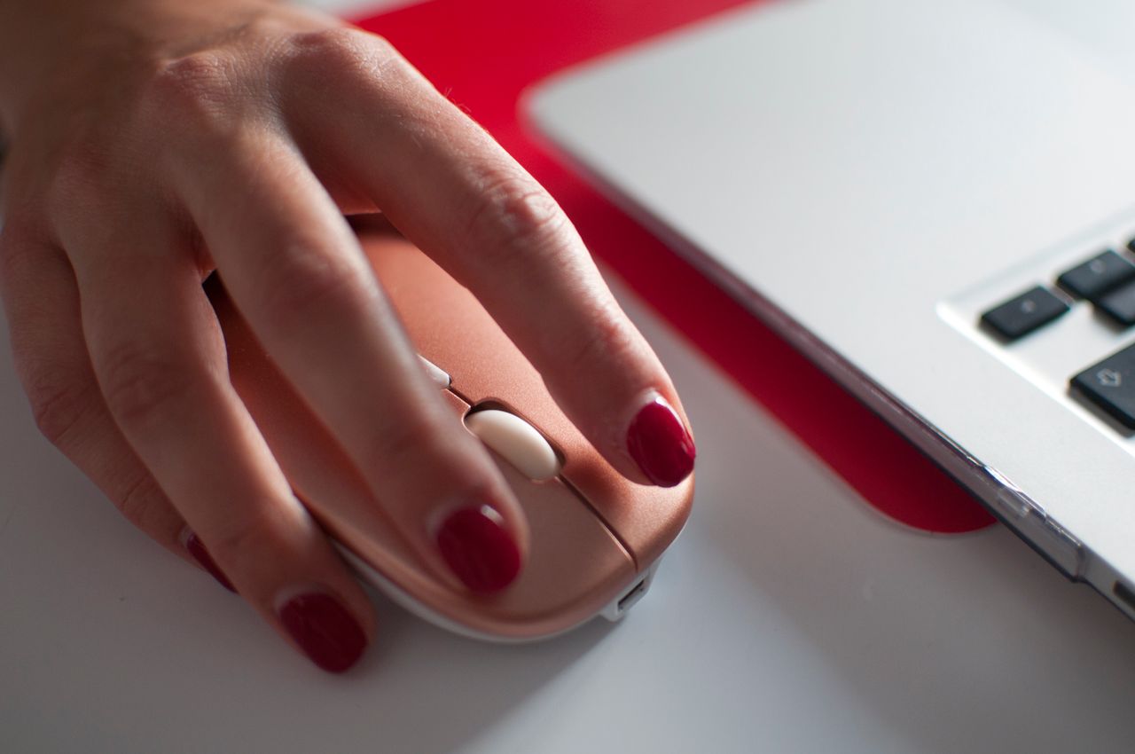 A womans hand with painted nails using a stylish computer mouse beside a laptop.